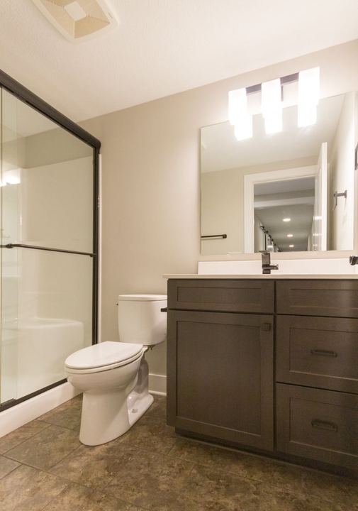 Basement bathroom with mottled brown stone-look tile floor, dark vanity and glass shower enclosure