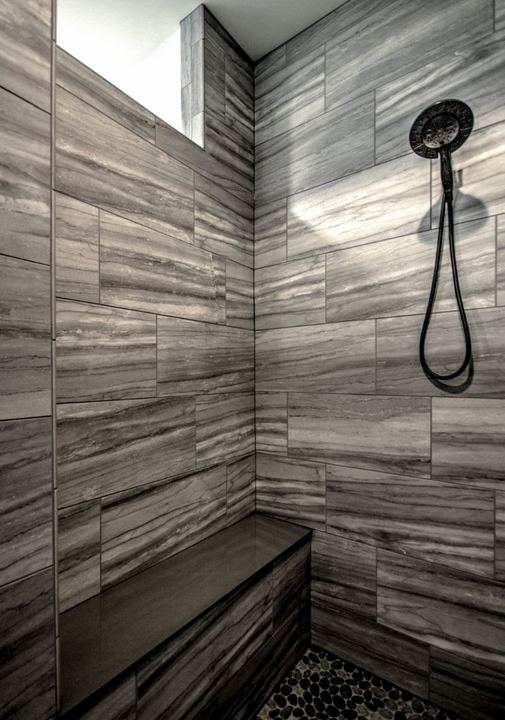 Walk-in shower with gray linear stone-look tile, tiled bench and black pebble floor