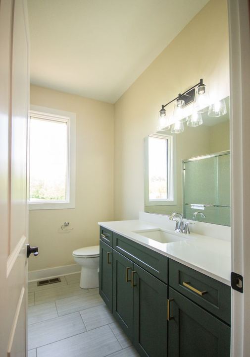 Hall bathroom with gray plank-look tile floor, green shaker vanity, brass pulls and white quartz top