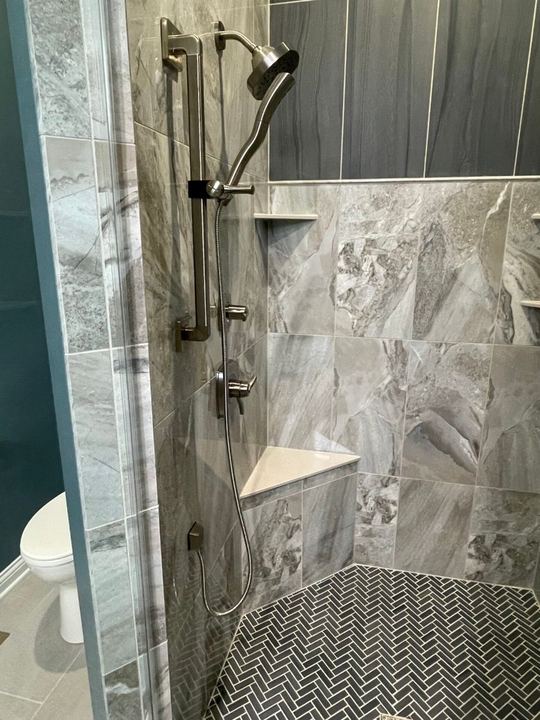 Master shower with gray marble-look tile walls, corner bench, herringbone mosaic floor and slide-bar handheld