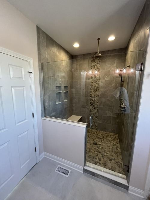 Walk-in shower with gray stone-look tile, vertical pebble mosaic accent stripe and pebble floor behind glass