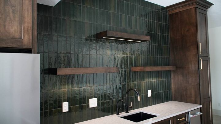Wet bar with glossy green vertical-stacked tile wall, walnut floating shelves, quartz counter and black faucet